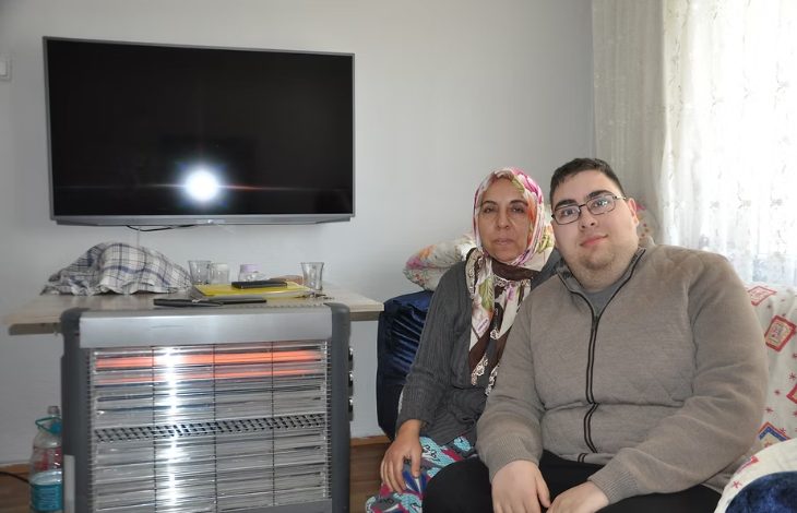 Fotoğrafta bir ev ortamında yan yana oturan bir anne ve oğlu görülüyor. Arkalarında duvara monte edilmiş bir televizyon ve önlerinde elektrikli ısıtıcı bulunuyor. İkili kameraya bakarak birlikte poz veriyor.