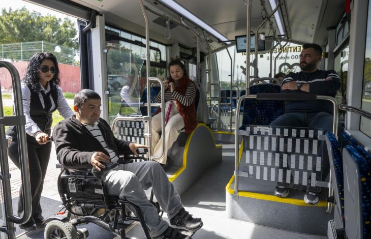 Kadın personel, tekerlekli sandalyedeki erkek yolcuyu otobüsün içine doğru yönlendirirken, içeride diğer özel gereksinimli yolcular koltuklarda oturuyor.