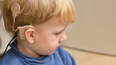 Fotoğrafta sarı saçlı küçük bir çocuk yan profilden görülüyor. Çocuğun kulağında işitme kaybı için kullanılan bir koklear implant cihazı bulunuyor. Cihaz kulağın arkasına yerleştirilmiş ve ince bir kabloyla kulak üstündeki parçaya bağlanmış. Çocuk mavi bir kazak giymiş ve dikkatle bir yere doğru bakıyor. Arka plan ise sade ve açık renkli bir iç mekân.