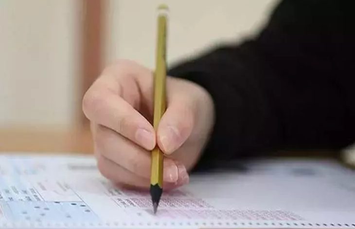 Görselde, bir kişinin sınav optik formu üzerinde kurşun kalemle işaretleme yaptığı yakın plan bir çekim görülüyor. Kalem, cevap alanına doğru tutulmuş ve odak formun üzerindeki işaretleme sürecinde.