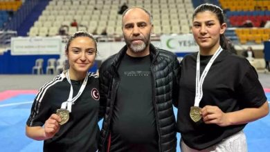 Bir spor salonunda çekilmiş fotoğrafta, ortada bir erkek ve iki yanında spor kıyafetli iki kadın yer alıyor. Kadın sporcular boyunlarında madalya taşıyor ve kameraya gülümseyerek poz veriyor. Arka planda tribünler ve minderli bir müsabaka alanı görülüyor.