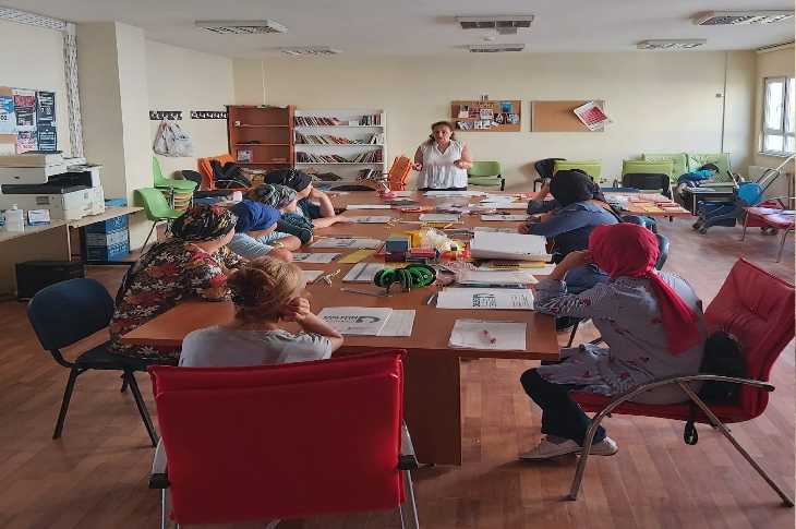 Görselde, geniş bir sınıf ortamında büyük bir masanın etrafına oturmuş yetişkin katılımcılar görülüyor. Masada defterler, çalışma kâğıtları ve çeşitli eğitim materyalleri bulunuyor. Odanın önünde ayakta duran bir eğitmen, grubun dikkatini kendisine vererek bir anlatım yapıyor. Arka planda kitaplıklar, panolar ve çalışma alanına ait mobilyalar yer alıyor.