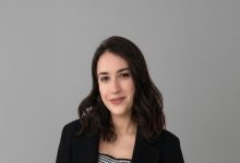 Kısa, sade bir arka plan önünde poz veren bir kadın görülüyor. Omuz hizasında dalgalı koyu renk saçlara sahip, siyah bir ceket ve desenli bir üst giymiş. Hafif bir gülümsemeyle kameraya bakıyor.