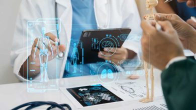 Bu görselde, bir sağlık çalışanı tablet kullanarak dijital bir sistem üzerinden insan iskeleti ve organlarını incelemekte, aynı zamanda masanın üzerinde küçük bir iskelet modeli ile insan anatomisi hakkında açıklama yapılmaktadır.