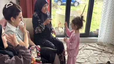 Bir oturma odasında üç yetişkin kadın ve küçük bir çocuk birlikte vakit geçiriyor. Yaşlı bir kadın, karşısında duran küçük kızla el hareketleriyle iletişim kurarken gülümsüyor. Yanındaki kadınlar da bu anı izleyip duygulanmış şekilde tepki veriyor. Arka planda büyük bir cam kapıdan dışarıda park edilmiş bir araba ve yeşil alan görünüyor. Ortam sıcak, samimi ve aile içi bir bayram buluşması hissi veriyor.