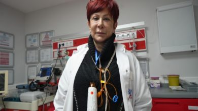 Görselde, bir sağlık kuruluşunun acil servis biriminde görevli bir sağlık çalışanı yer alıyor. Beyaz önlük giymiş olan kişi, üzerinde çeşitli tıbbi ekipmanlar taşıyor. Arka planda monitörler, tıbbi cihazlar ve acil müdahale malzemelerinin bulunduğu bir çalışma alanı görünüyor. Atmosfer, yoğun bir sağlık hizmeti ortamını yansıtıyor.