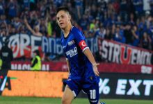 Bir futbol maçında, mavi formalı bir oyuncu sahada koşarken görülüyor. Kolunda kaptanlık bandı var ve üzerinde Türk bayrağı detayı dikkat çekiyor. Tribünler dolu, arka planda coşkulu taraftarlar ve pankartlar yer alıyor. Oyuncu gol ya da önemli bir pozisyon sonrası sevinç anı yaşıyor gibi görünüyor.