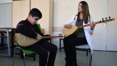 Bir sınıf ortamında, sandalyede oturan bir çocuk bağlama çalmaya çalışırken yanında oturan bir kadın da bağlama çalarak ona eşlik ediyor. Kadın çocuğa rehberlik eder gibi dikkatle bakarken, çocuk enstrümanına odaklanmış durumda. Arka planda dolaplar ve sınıf eşyaları görülüyor.