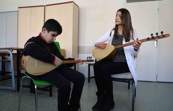 Bir sınıf ortamında, sandalyede oturan bir çocuk bağlama çalmaya çalışırken yanında oturan bir kadın da bağlama çalarak ona eşlik ediyor. Kadın çocuğa rehberlik eder gibi dikkatle bakarken, çocuk enstrümanına odaklanmış durumda. Arka planda dolaplar ve sınıf eşyaları görülüyor.