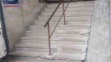 Beton duvarlar arasında yer alan, ortasından paslı bir metal korkuluk geçen ve kenarları aşınmış taş basamaklardan oluşan eski bir merdiven ile dik yapılmış engelli rampası görülüyor.