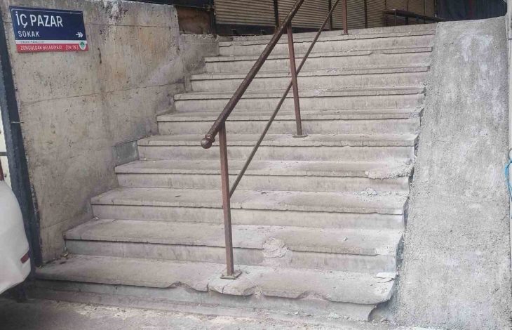 Beton duvarlar arasında yer alan, ortasından paslı bir metal korkuluk geçen ve kenarları aşınmış taş basamaklardan oluşan eski bir merdiven ile dik yapılmış engelli rampası görülüyor.