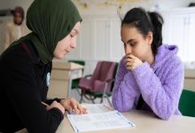 Görme engelli kız öğrenciye, akranı kız öğrenci soru kitapçığı okuyor.