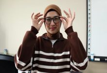 Genç bir kadın, kahverengi tonlarda çizgili bir üst ve başörtüsüyle sınıf benzeri bir ortamda duruyor. Yuvarlak çerçeveli gözlüğünü iki eliyle düzeltiyor ve hafifçe gülümsüyor. Arka planda bir beyaz tahta ve eğitim materyalleri görülüyor.