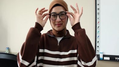 Genç bir kadın, kahverengi tonlarda çizgili bir üst ve başörtüsüyle sınıf benzeri bir ortamda duruyor. Yuvarlak çerçeveli gözlüğünü iki eliyle düzeltiyor ve hafifçe gülümsüyor. Arka planda bir beyaz tahta ve eğitim materyalleri görülüyor.