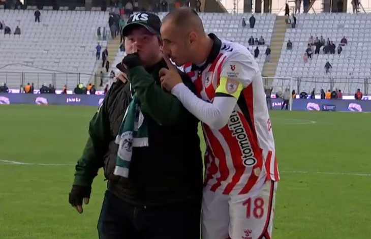 Stadyumda, bir futbolcu forma ve kaptan bandıyla sahada dururken, yanında bulunan taraftarın kulağına eğilip samimi bir şekilde konuşuyor. Taraftar atkı ve şapka takmış; arka planda tribünler ve saha atmosferi görülüyor.