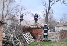 Görselde, bir yapının çatısında çıkan yangına müdahale eden itfaiye ekipleri görülüyor. Dumanın yoğun olduğu alanda üç itfaiyeci hortum ve ekipmanlarıyla söndürme çalışması yürütüyor. Yapı kısmen yıkılmış halde; çevrede odun yığınları, duvar kalıntıları ve dağınık malzemeler bulunuyor. Atmosfer, acil müdahalenin sürdüğünü gösteriyor.