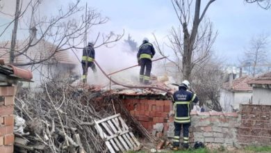 Görselde, bir yapının çatısında çıkan yangına müdahale eden itfaiye ekipleri görülüyor. Dumanın yoğun olduğu alanda üç itfaiyeci hortum ve ekipmanlarıyla söndürme çalışması yürütüyor. Yapı kısmen yıkılmış halde; çevrede odun yığınları, duvar kalıntıları ve dağınık malzemeler bulunuyor. Atmosfer, acil müdahalenin sürdüğünü gösteriyor.
