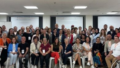 Geniş bir salonda, farklı yaş gruplarından kalabalık bir ekip toplu fotoğraf için bir araya gelmiş. Ön sırada sandalyede oturanlar, arkada ayakta duranlarla birlikte kameraya gülümserken çoğunun boynunda etkinlik kartları bulunuyor. Ortam, bir zirve ya da toplantı sonrası çekilmiş hatıra fotoğrafı havası taşıyor.