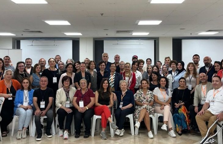 Geniş bir salonda, farklı yaş gruplarından kalabalık bir ekip toplu fotoğraf için bir araya gelmiş. Ön sırada sandalyede oturanlar, arkada ayakta duranlarla birlikte kameraya gülümserken çoğunun boynunda etkinlik kartları bulunuyor. Ortam, bir zirve ya da toplantı sonrası çekilmiş hatıra fotoğrafı havası taşıyor.