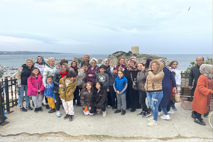 Deniz manzaralı bir noktada, aralarında çocukların da bulunduğu kalabalık bir grup birlikte fotoğraf çektiriyor. Arka planda deniz, kıyı yerleşimi ve küçük bir tarihi yapı görülüyor. Grup samimi ve neşeli bir atmosfer içinde bir araya gelmiş.