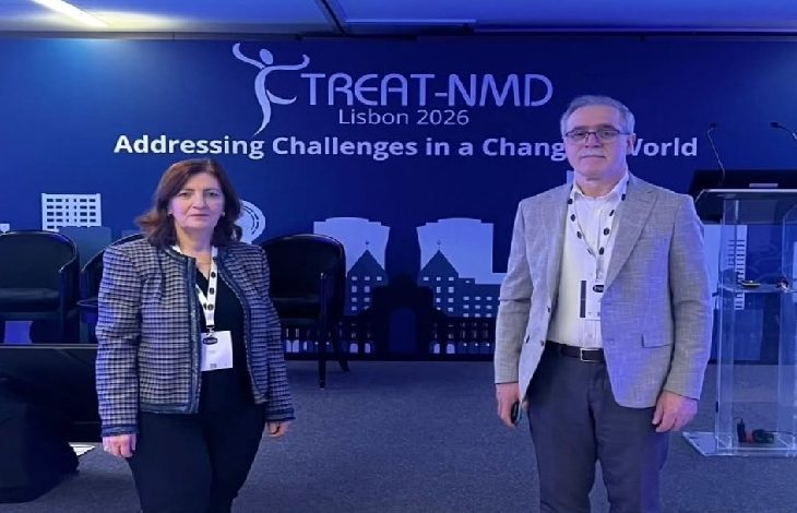 Görselde, bir konferans salonunda sahne önünde duran iki kişi görülüyor. Arkadaki büyük ekranda “TREAT-NMD Lisbon 2026 – Addressing Challenges in a Changing World” yazısı yer alıyor. Kadın ve erkek katılımcılar yaka kartlarıyla kamera karşısında poz verirken, sahnede panel koltukları ve konuşma kürsüsü de dikkat çekiyor. Bu kare, uluslararası bir konferans etkinliği sırasında çekilmiş gibi görünüyor.