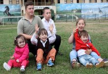 Çim alanda bir araya gelmiş, çoğu Down sendromlu olan çocuklar ve iki genç oturmuş poz veriyor. Bazıları gülüyor, bazıları ellerini kaldırmış; arka planda ise duvar resimleriyle süslenmiş bir okul bahçesi görünüyor.