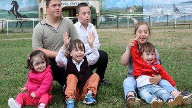 Çim alanda bir araya gelmiş, çoğu Down sendromlu olan çocuklar ve iki genç oturmuş poz veriyor. Bazıları gülüyor, bazıları ellerini kaldırmış; arka planda ise duvar resimleriyle süslenmiş bir okul bahçesi görünüyor.