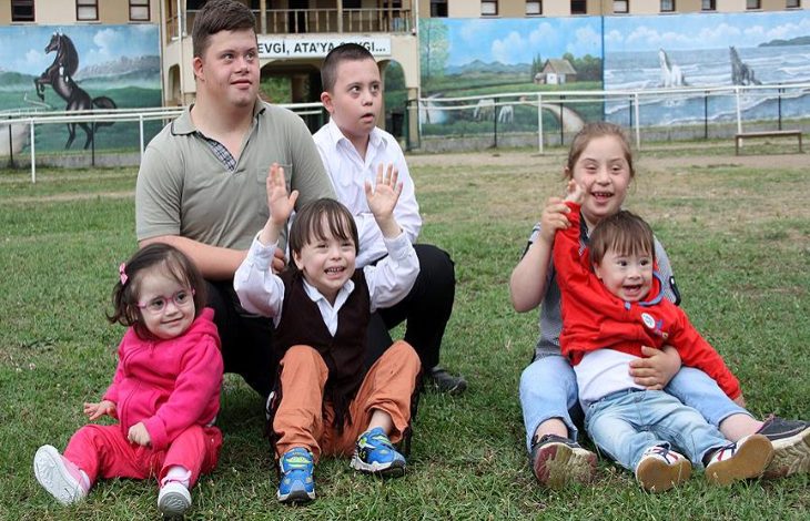 Çim alanda bir araya gelmiş, çoğu Down sendromlu olan çocuklar ve iki genç oturmuş poz veriyor. Bazıları gülüyor, bazıları ellerini kaldırmış; arka planda ise duvar resimleriyle süslenmiş bir okul bahçesi görünüyor.