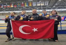 Kapalı bir spor salonunda çekilmiş fotoğrafta, “Türkiye” yazılı spor kıyafetleri giyen dört kadın sporcu ve ortalarında duran bir erkek görülüyor. Sporcular boyunlarında altın madalyalarını gösterirken birlikte büyük bir Türk bayrağı tutuyor. Arka planda pist alanı ve farklı ülkelere ait bayraklar yer alıyor. Görsel, uluslararası bir spor organizasyonunda kazanılan başarıyı yansıtan bir anı gösteriyor.