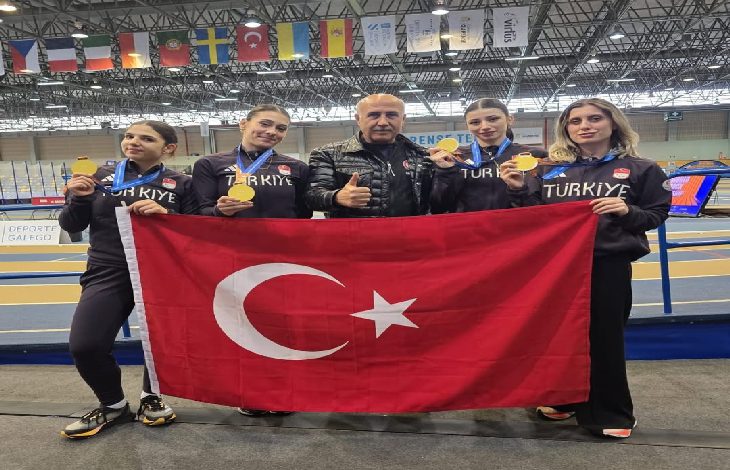 Kapalı bir spor salonunda çekilmiş fotoğrafta, “Türkiye” yazılı spor kıyafetleri giyen dört kadın sporcu ve ortalarında duran bir erkek görülüyor. Sporcular boyunlarında altın madalyalarını gösterirken birlikte büyük bir Türk bayrağı tutuyor. Arka planda pist alanı ve farklı ülkelere ait bayraklar yer alıyor. Görsel, uluslararası bir spor organizasyonunda kazanılan başarıyı yansıtan bir anı gösteriyor.