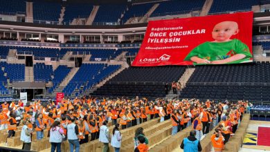 Spor salonunda, turuncu yelekli çok sayıda gönüllü uzun sıralar halinde dizilmiş kolilerin arasında çalışırken, tribünlere asılmış büyük kırmızı pankartta “Bağışlarınızla önce çocuklar iyileşsin” yazısı ve bir bebeğin fotoğrafı yer alıyor.