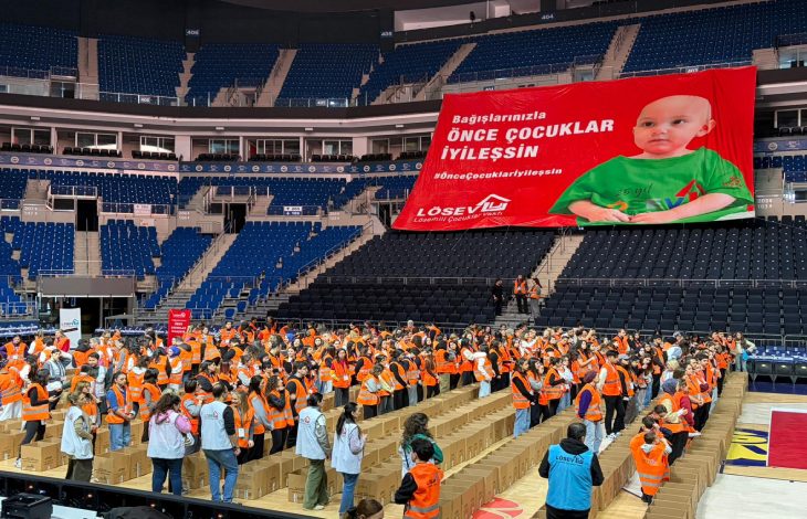 Spor salonunda, turuncu yelekli çok sayıda gönüllü uzun sıralar halinde dizilmiş kolilerin arasında çalışırken, tribünlere asılmış büyük kırmızı pankartta “Bağışlarınızla önce çocuklar iyileşsin” yazısı ve bir bebeğin fotoğrafı yer alıyor.