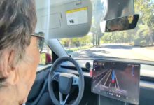 Direksiyon başında yaşlı bir kadın görülüyor. Araç içinde büyük bir dijital ekran var ve yolda ilerlerken sürüş destek sistemi açık görünüyor. Dışarıda ağaçlık bir yol uzanıyor.