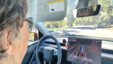 Direksiyon başında yaşlı bir kadın görülüyor. Araç içinde büyük bir dijital ekran var ve yolda ilerlerken sürüş destek sistemi açık görünüyor. Dışarıda ağaçlık bir yol uzanıyor.