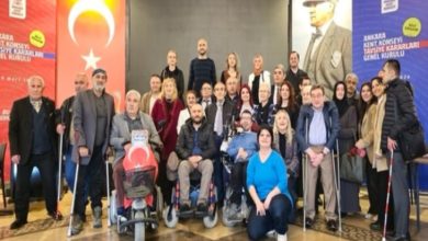 Kalabalık bir grup, kapalı bir salonda toplu fotoğraf için poz veriyor. Aralarında tekerlekli sandalye kullananlar ve bastonla ayakta duranlar var. Arkada Türk bayrağı ve Mustafa Kemal Atatürk’ün portresi yer alıyor. Katılımcılar resmi bir etkinlik sonrası bir araya gelmiş gibi görünüyor.