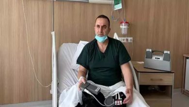 Hastane odasında yatağa oturmuş bir adam görülüyor. Boynunda maskesi var, sakin ama yorgun bir ifadeyle kameraya bakıyor. Kucağında ve yanında kablolarla bağlı taşınabilir bir tıbbi cihaz bulunuyor; bu cihazla yaşamını sürdürüyor gibi görünüyor. Etrafında serum askısı ve tıbbi ekipmanlar yer alıyor.