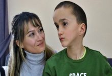 Bir odada çekilmiş fotoğrafta, kısa saçlı küçük bir çocuk yana doğru bakarken, yanında oturan bir kadın ona sevgi ve dikkatle bakıyor. Kadının yüzünde hafif bir tebessüm var; aralarındaki bağ sıcak ve koruyucu bir his veriyor. Çocuk ise düşünceli ve biraz dalgın görünüyor.