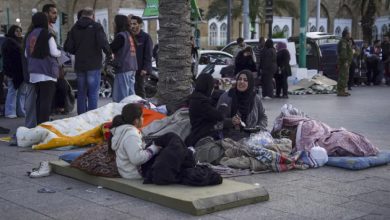Açık alanda, yerde serili yatak ve battaniyeler üzerinde oturan bir aile görülüyor. Kadınlar ve çocuklar sokakta kalmış; etrafta eşyalar dağınık. Arka planda kalabalık insanlar ve hareketli bir şehir ortamı dikkat çekiyor.