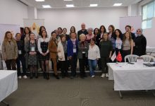 Görselde, bir salon ortamında düzenlenen etkinlik sonrası toplu halde poz veren kalabalık bir grup görülüyor. Çoğunluğu kadınlardan oluşan katılımcılar, sahnenin önünde yan yana durarak kameraya gülümsüyor. Arka planda etkinliğe ait afişler ve masaların bulunduğu bir çalışma alanı yer alıyor. Ortam, profesyonel ve iş birliğine dayalı bir toplantı atmosferi yansıtıyor.