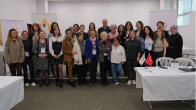 Görselde, bir salon ortamında düzenlenen etkinlik sonrası toplu halde poz veren kalabalık bir grup görülüyor. Çoğunluğu kadınlardan oluşan katılımcılar, sahnenin önünde yan yana durarak kameraya gülümsüyor. Arka planda etkinliğe ait afişler ve masaların bulunduğu bir çalışma alanı yer alıyor. Ortam, profesyonel ve iş birliğine dayalı bir toplantı atmosferi yansıtıyor.