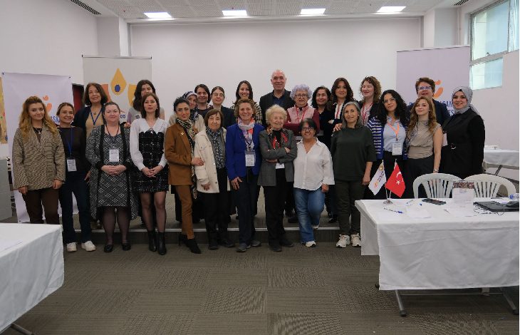 Görselde, bir salon ortamında düzenlenen etkinlik sonrası toplu halde poz veren kalabalık bir grup görülüyor. Çoğunluğu kadınlardan oluşan katılımcılar, sahnenin önünde yan yana durarak kameraya gülümsüyor. Arka planda etkinliğe ait afişler ve masaların bulunduğu bir çalışma alanı yer alıyor. Ortam, profesyonel ve iş birliğine dayalı bir toplantı atmosferi yansıtıyor.