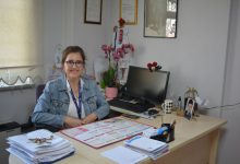 Görselde, bir ofis ortamında masasında oturan ve gülümseyen bir kadın görülüyor. Masanın üzerinde takvim, belgeler, kalemlik, bilgisayar ve çeşitli kişisel eşyalar yer alıyor. Arka planda çerçeveli belgeler ve çiçekler bulunuyor. Ortam sakin ve düzenli bir çalışma alanı izlenimi veriyor.