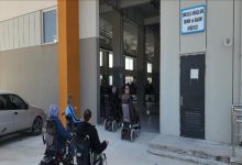 Tekerlekli sandalye kullanan bir grup bireyin, üzerinde "Engelli Araçları Tamir ve Bakım Atölyesi" tabelası bulunan bir binaya giriş yaptığı anı gösteren görsel. Fotoğraf, engelli bireylerin hareketliliğini destekleyen bir hizmet merkezini ve bu merkeze olan erişimi yansıtıyor.