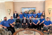 Bir oturma salonunda, aralarında tekerlekli sandalye kullanan sporcuların da bulunduğu kalabalık bir ekip toplu fotoğraf veriyor. Ortada yaşça büyük iki kişi otururken, etraflarında mavi formalarıyla takım üyeleri yer alıyor; arka planda tablo ve klasik dekor dikkat çekiyor.