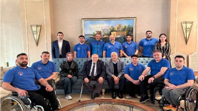 Bir oturma salonunda, aralarında tekerlekli sandalye kullanan sporcuların da bulunduğu kalabalık bir ekip toplu fotoğraf veriyor. Ortada yaşça büyük iki kişi otururken, etraflarında mavi formalarıyla takım üyeleri yer alıyor; arka planda tablo ve klasik dekor dikkat çekiyor.