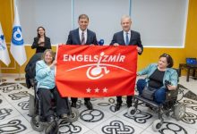 Görselde, İzmir Büyükşehir Belediye Başkanı Dr. Cemil Tugay, İZTO Yönetim Kurulu Başkanı Mahmut Özgener ile iki tekerlekli sandalye kullanan kadının birlikte “Engelsiz İzmir” yazılı kırmızı bir pankart tutarak engelli bireyler için farkındalık mesajı verdiği bir etkinlik anı görülüyor.