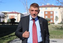 Açık havada çekilmiş bir portre görülüyor. Takım elbise ve kırmızı çizgili kravat takan orta yaşlı bir erkek, yürüyüş yolunda kameraya doğru ilerlerken ceketini tutuyor. Arka planda apartmanlar, ağaçlar ve güneşli bir günün net ışığı var. Yüz ifadesi ciddi ama kararlı bir duruş yansıtıyor.