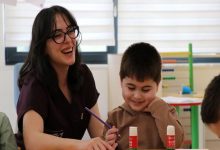 Bir sınıf ortamında, gözlüklü bir kadın öğretmen küçük bir çocukla birlikte masa başında etkinlik yapıyor. Kadın gülerek çocuğa eşlik ederken, çocuk elinde kalemle çalışıyor ve mutlu görünüyor. Masada yapıştırıcılar var; arka planda renkli eğitim materyalleri ve başka bir çocuk daha kısmen görünüyor. Atmosfer sıcak ve destekleyici bir öğrenme anını yansıtıyor.