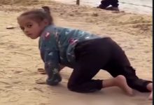 Kumlu bir zeminde, küçük bir çocuk emekler halde ilerliyor. Başını geriye çevirip kameraya bakıyor. Üzerinde uzun kollu bir üst ve koyu renk pantolon var; arka planda bulanık şekilde başka biri görülüyor.