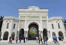 Geniş bir meydanda yer alan tarihi ve anıtsal bir üniversite kapısı görülüyor. Üzerinde süslemeler, kitabeler ve saat kuleleri bulunan yapı oldukça görkemli. Kapının önünde farklı yönlere yürüyen öğrenciler var; bazıları sohbet ederken bazıları tek başına ilerliyor. Hava açık ve mekân canlı bir kampüs atmosferi taşıyor.